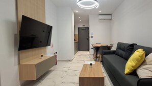 A modern living room with a flat-screen TV mounted on the wall, a wooden coffee table, a black sofa with yellow and beige cushions, and a small table with chairs in the background. - Ilion Apartments (Thessaloniki)