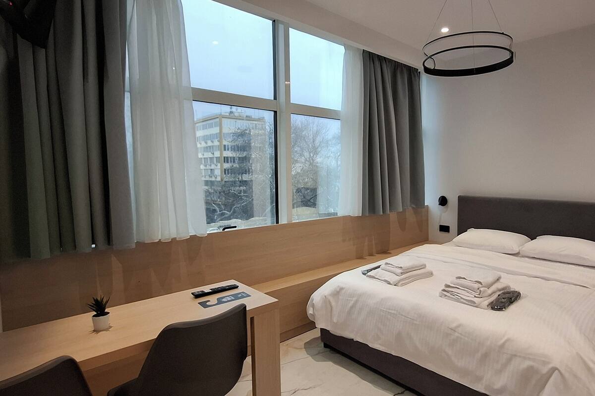 A modern hotel room with a large bed, a desk, and a view of a building through the window.