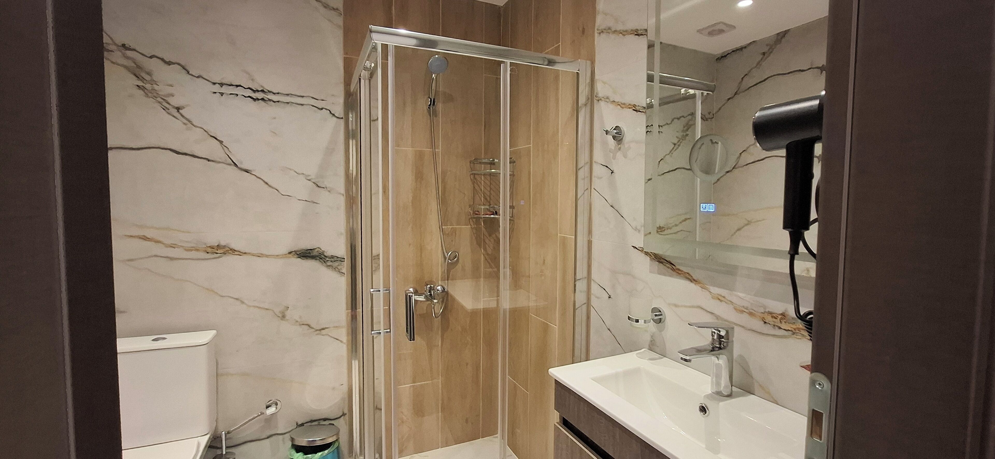 Un bagno moderno con doccia vetrata, parete rivestita in marmo, lavandino bianco e phon sul piano di appoggio.