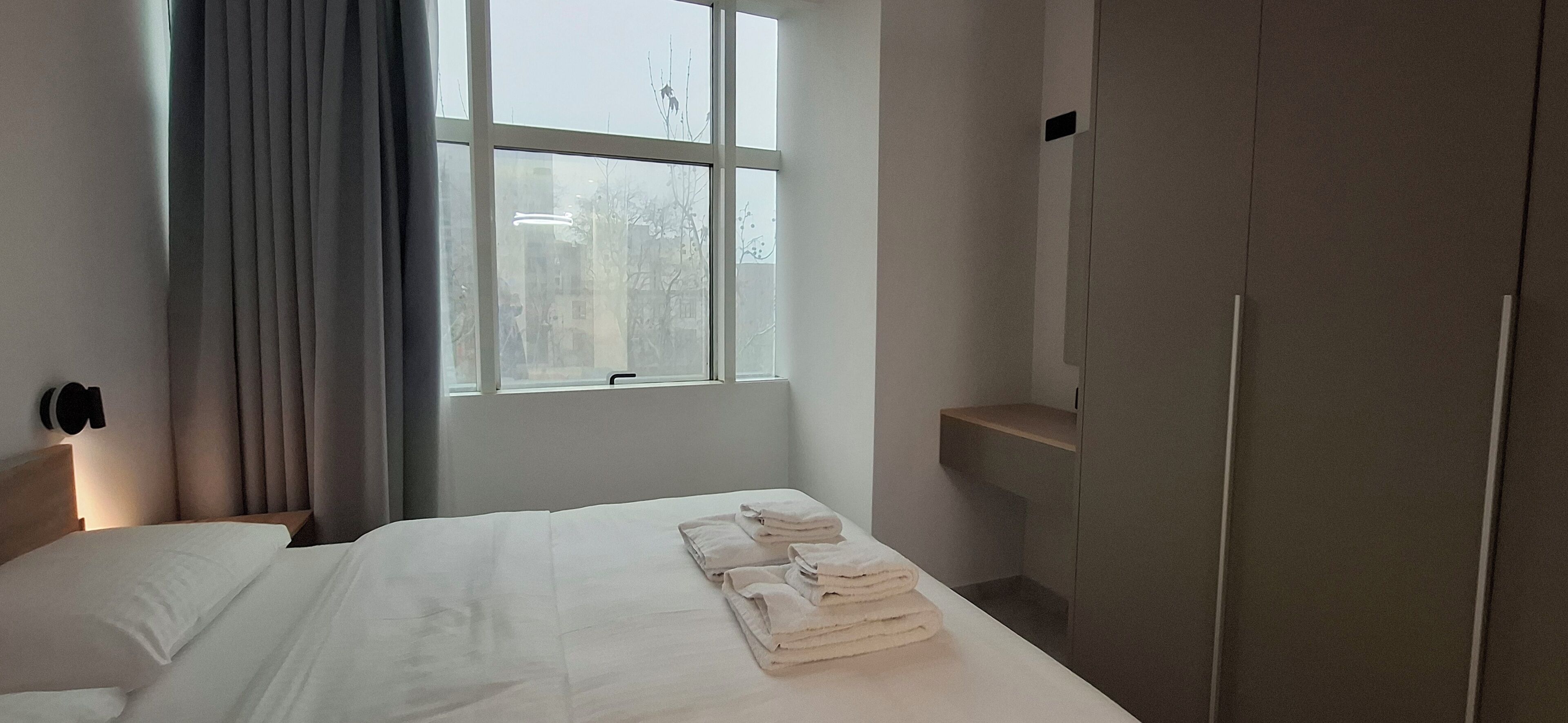 A hotel room with a bed, a window with curtains, a wardrobe, and a wall-mounted light.