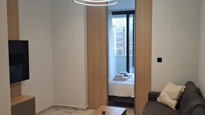 Moderni hotellihuone, jossa on taulutelevisio, sohva, pieni pöytä ja sänky, jossa on siististi viikatut valkoiset vuodevaatteet.