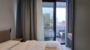 A hotel room with a bed, a small table, and a view of the city through a glass door. - Ilion Apartments (Thessaloniki)