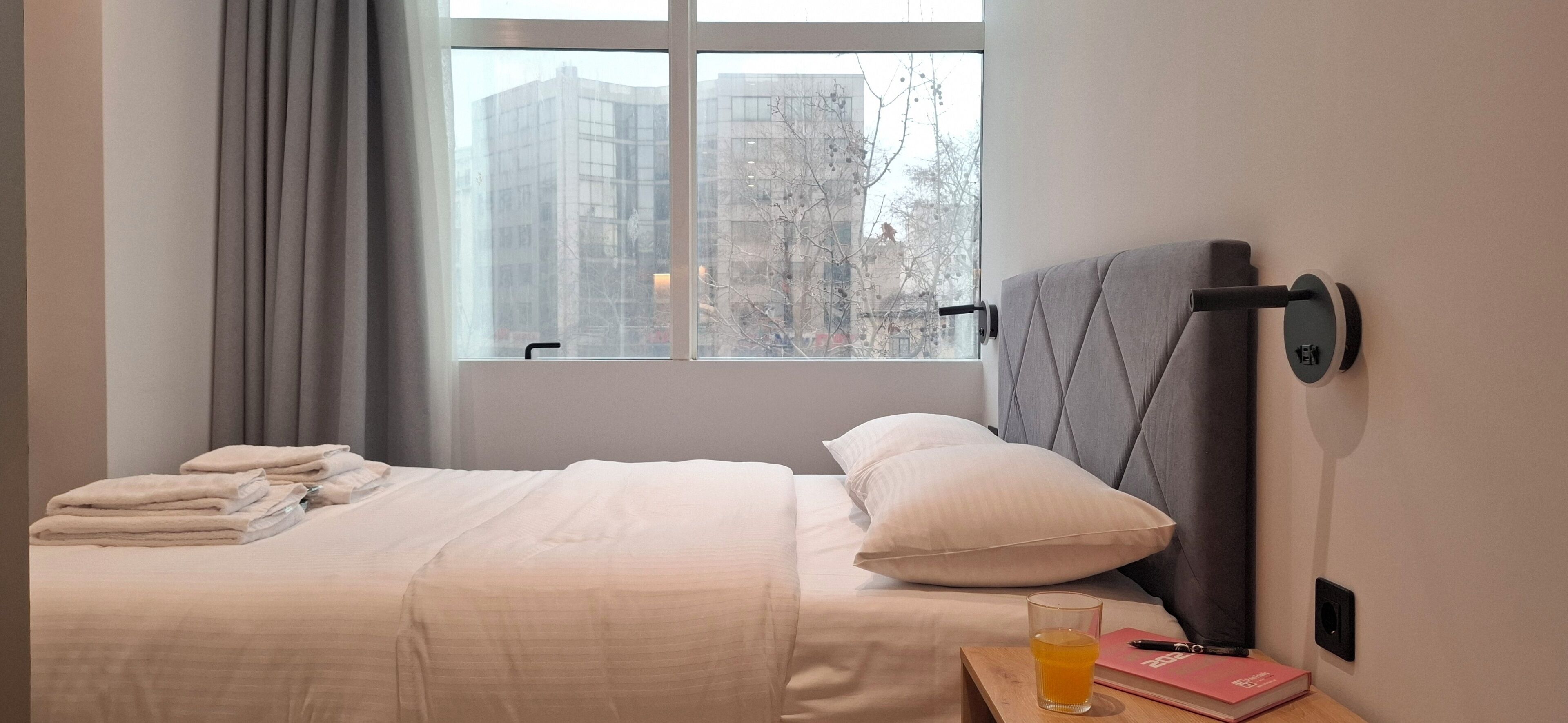 A neatly made bed with a headboard, a bedside table with a glass of orange juice and a pink book, and a window with a view of buildings and trees.