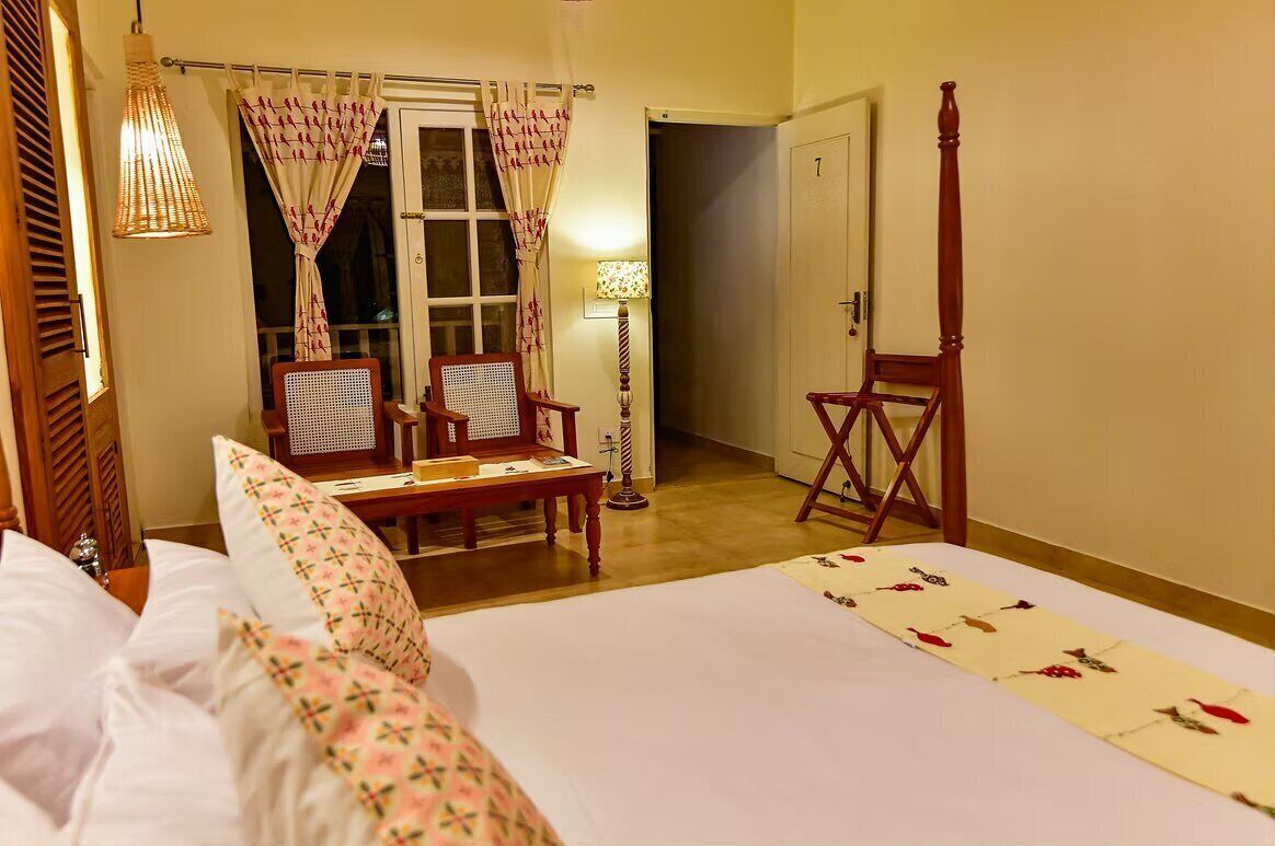 A hotel room with a bed, two chairs, a table, a lamp, and a wooden chair.