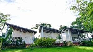 A row of three white buildings with a covered porch, surrounded by lush greenery and a well-maintained lawn. - Musafir Jungle Resort (Sarupeta)