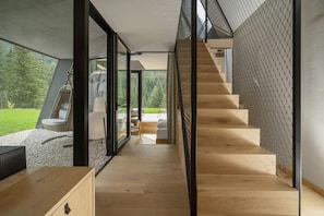 Un interior moderno con una escalera de madera, puertas de cristal que dan a un balcón y vistas a un paisaje verde.