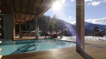 Eine Holzterrasse mit Pool, umgeben von verschneiten Bergen und blauem Himmel.