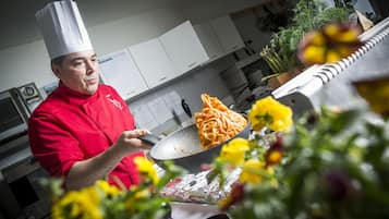 Ein Koch in einer roten Uniform bereitet Spaghetti in einer Küche mit weißen Schränken zu. Im Hintergrund ist eine Pflanze zu sehen.