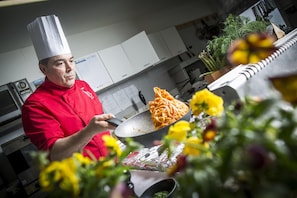 Un chef con uniforme rojo cocinando espagueti en una cocina con armarios blancos y una planta al fondo.