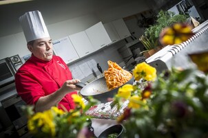 Un chef con uniforme rojo cocinando espagueti en una cocina con armarios blancos y una planta al fondo.