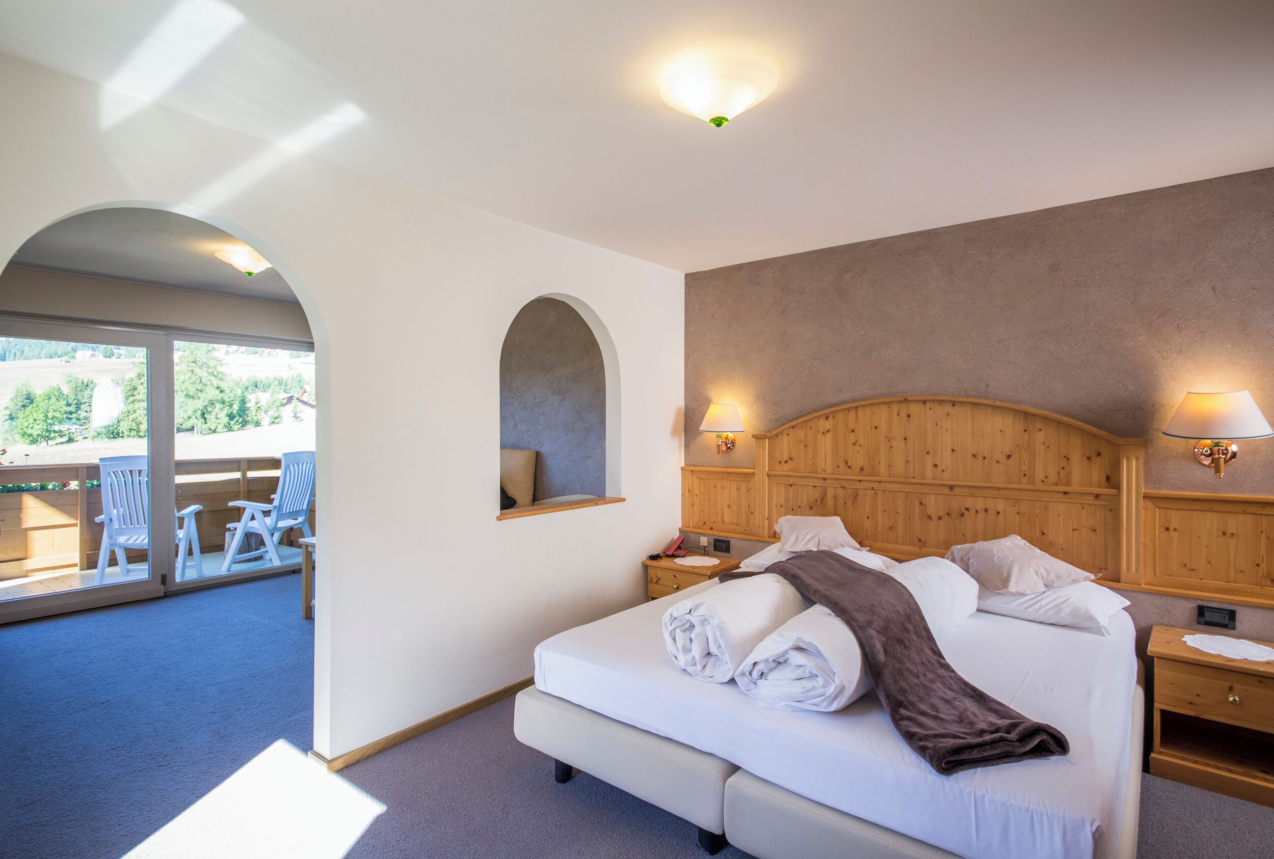 Un dormitorio con una cama grande, cabecera de madera, dos mesitas de noche y una ventana con vistas a una zona de estar exterior.