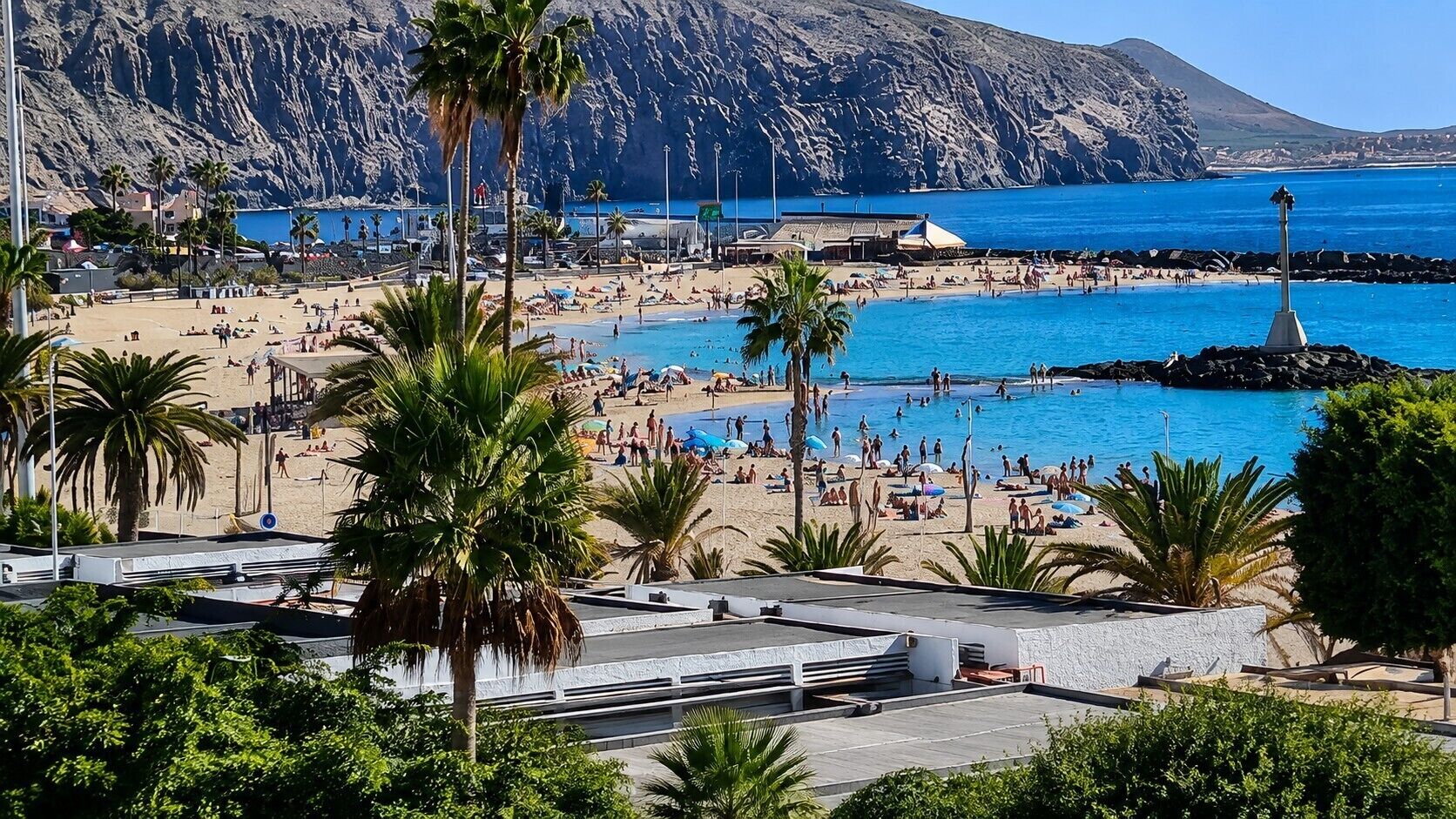 A beach with a pool, palm trees, and a rocky mountain in the background.