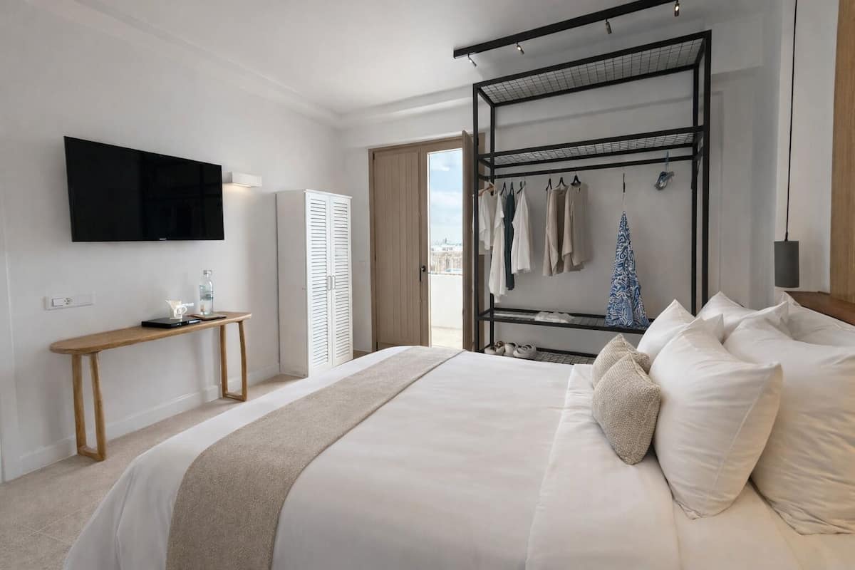 Habitació d'hotel moderna amb un llit gran, una televisió penjada a la paret, una taula de nit de fusta, una estant per a roba i vista a la ciutat per la finestra.