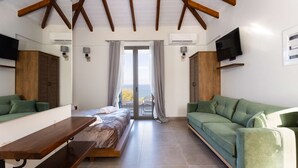 A room with a bed, a green sofa, a wooden bench, a TV, and a sliding glass door leading to an outdoor area. - Eneos Boutique Residence - Koroni Garden Jewel (Pylos-Nestoras)