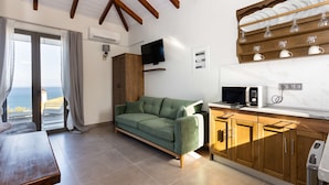 A modern living room with a green sofa, wooden cabinets, a flat-screen TV, and a kitchen area with a microwave. - Eneos Boutique Residence - Koroni Garden Jewel (Pylos-Nestoras)
