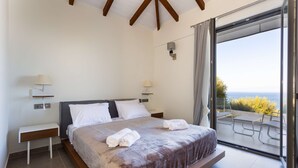 A bedroom with a large bed, two bedside tables with lamps, a sliding glass door leading to an outdoor area with a table and chairs. - Eneos Boutique Residence - Koroni Garden Jewel (Pylos-Nestoras)