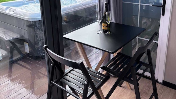 A glass-enclosed patio with a table and chairs, a hot tub visible through the glass, and a view of a garden and house in the background. - Glamping Pod With Hot Tub in Orchard (Milford Haven)
