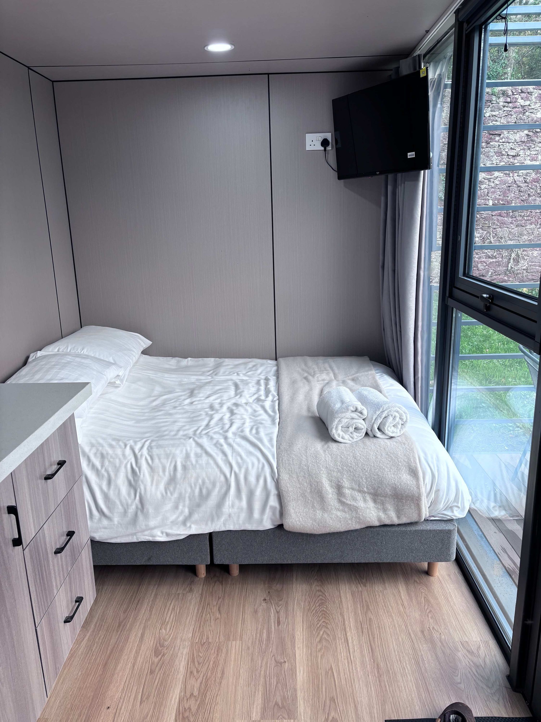 A compact bedroom with a bed, a bedside table, a TV mounted on the wall, and a window with a view of greenery.