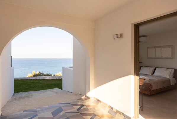 A bedroom with a bed, a nightstand, and a view of the sea through an arched doorway. - Eneos Boutique Residence - Koroni Garden Gem (Pylos-Nestoras)