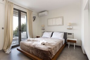 A modern bedroom with a large bed, two bedside tables, a wall-mounted air conditioner, and a sliding glass door leading to a balcony. - Eneos Boutique Residence - Koroni Garden Gem (Pylos-Nestoras)