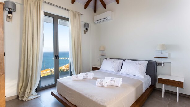 A bedroom with a large bed, a nightstand, a lamp, and a view of the sea. - Eneos Boutique Residence - Koroni Garden Nest (Pylos-Nestoras)
