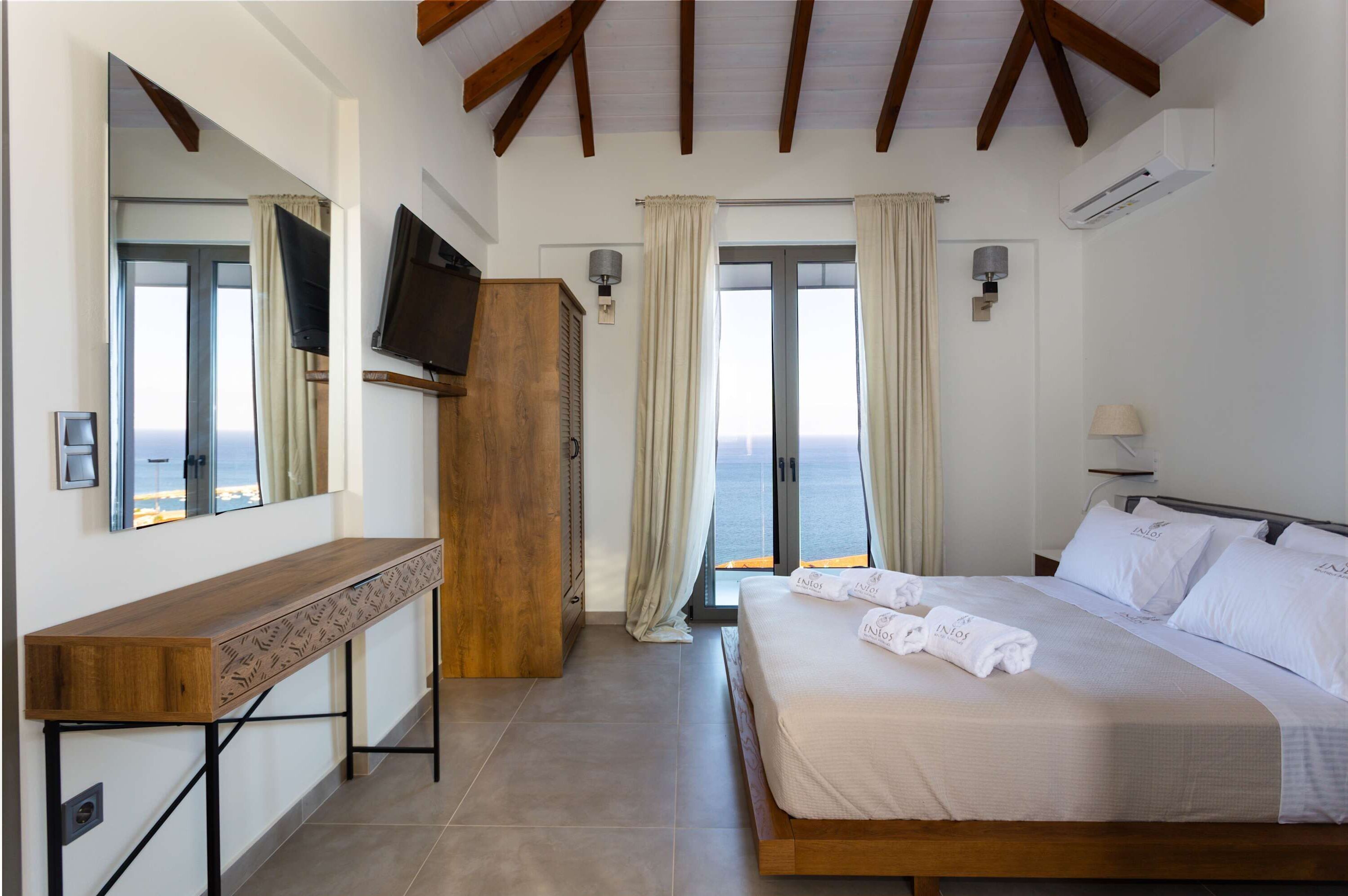 Une chambre à coucher avec un lit, un bureau en bois, une télévision et une vue sur la mer grâce à de grandes fenêtres.