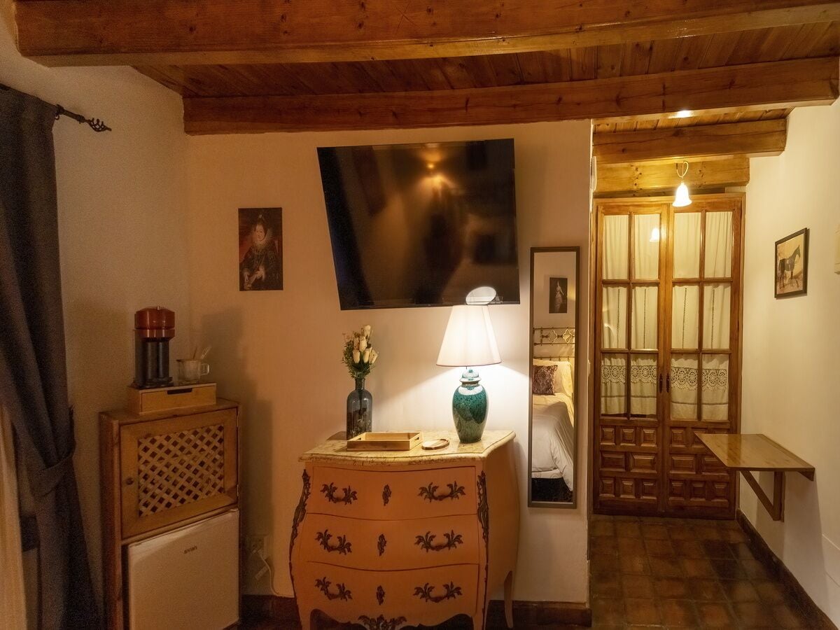A cozy room with a wooden ceiling, a small cabinet with a coffee maker, a vase with flowers, a lamp, and a door leading to another room.