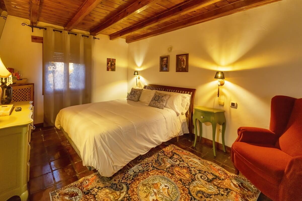 Une chambre confortable avec un plafond en bois, un grand lit avec une literie blanche, deux lampes fixées au mur, un tapis à motifs et un fauteuil rouge.