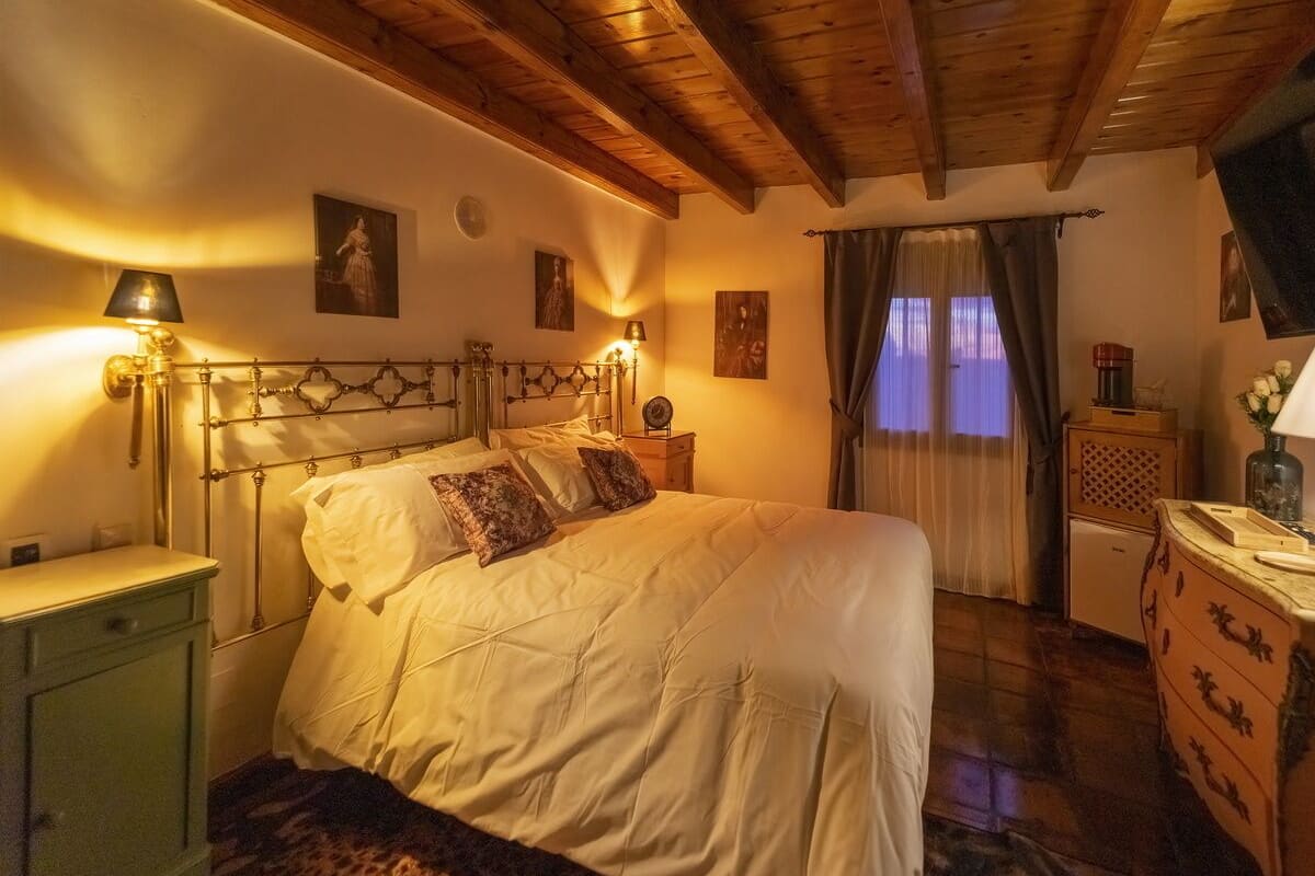 Une chambre confortable avec un plafond en bois, un lit avec une literie blanche, une table de chevet avec une lampe et une commode ornée d’un vase de fleurs.