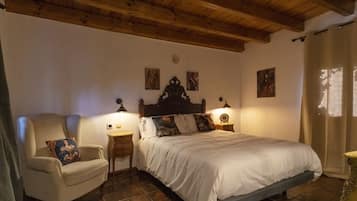 A bedroom with a wooden ceiling, a bed with white bedding, a chair with a cushion, two bedside tables with lamps, and a window with curtains.