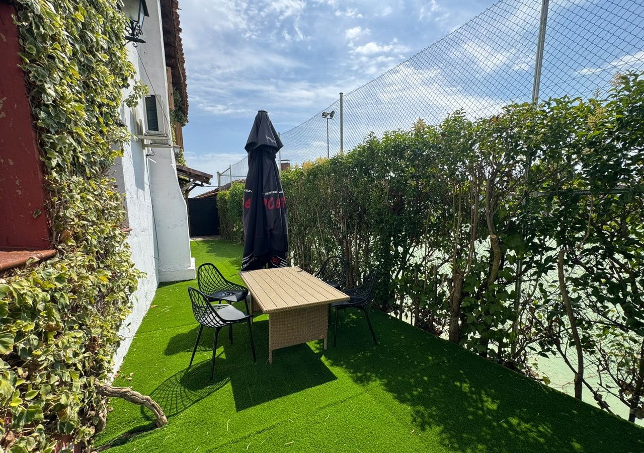 Un patio exterior con mesa y sillas, rodeado de vegetación y una cerca.