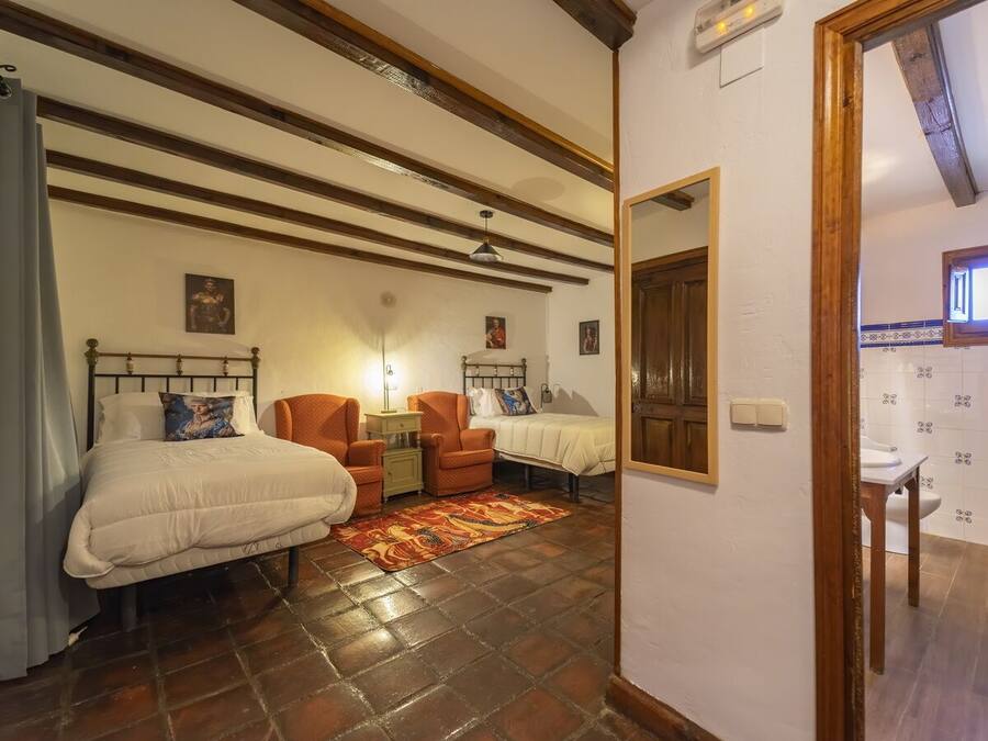 A bedroom with two beds, a nightstand, an orange armchair, a patterned rug, and a wooden ceiling with exposed beams.
