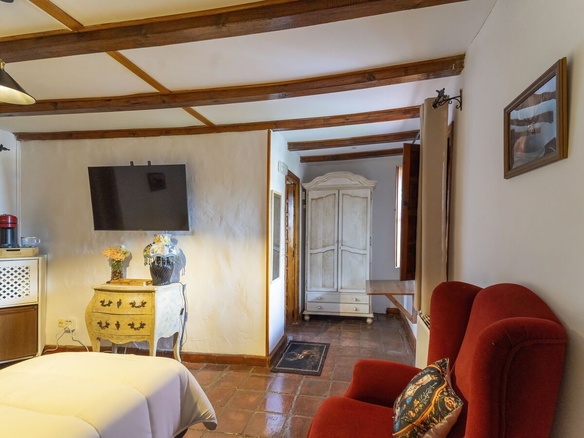 A room with a bed, a red armchair, a TV, a wardrobe, and a door leading to another room.