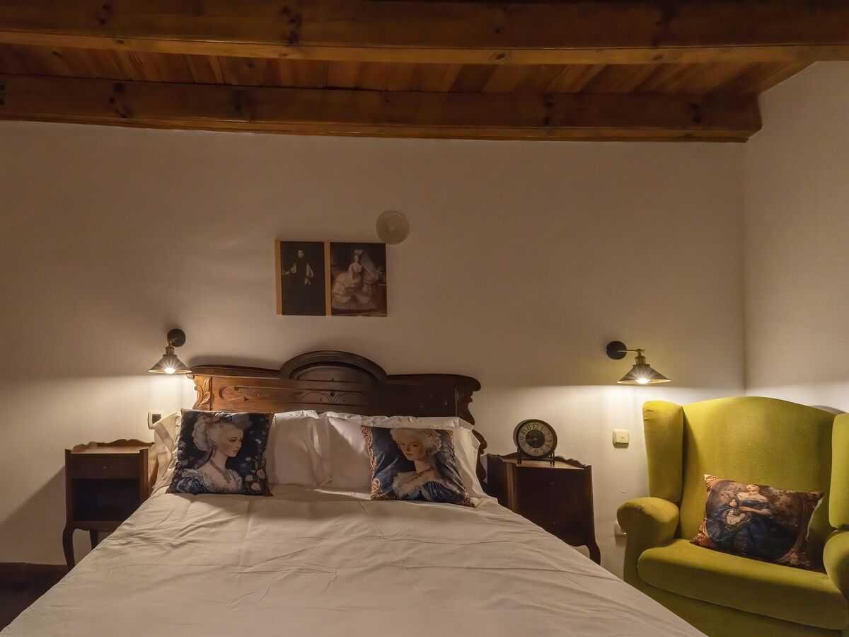 A bedroom with a wooden ceiling, a bed with decorative pillows, a green armchair, and two wall-mounted lamps.