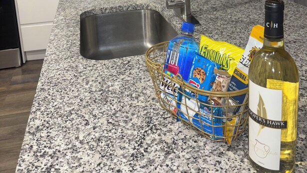 Une cuisine dotée d’un plan de travail en granit, d’un évier en acier inoxydable, d’un robinet, d’un panier rempli d’en-cas, d’une bouteille d’eau Fiji et d’une bouteille de vin.