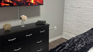 Une chambre avec une commode noire, un téléviseur diffusant une image de cheminée, un mur en briques blanches et un tapis noir.