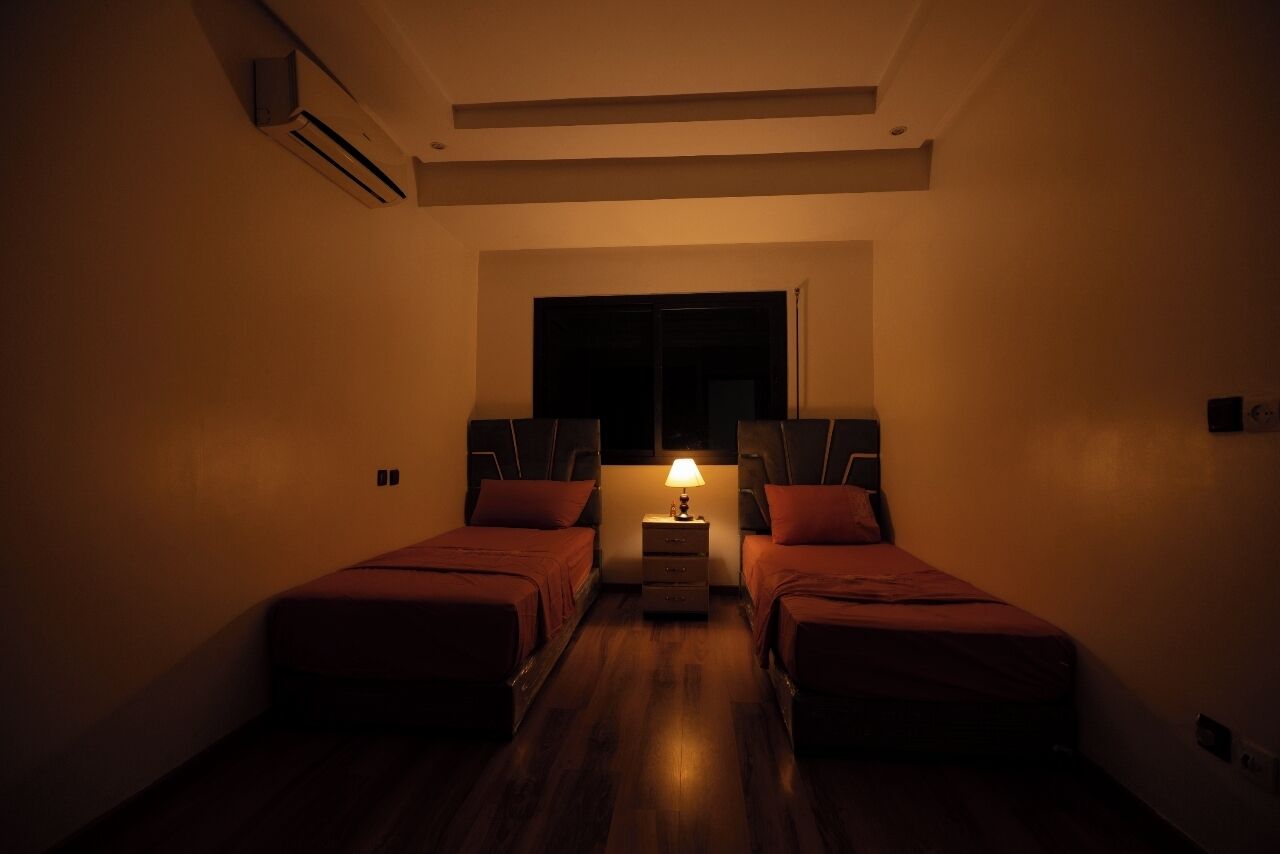 A dimly lit hotel room with two beds, a nightstand with a lamp, and a window with a view of the outside.
