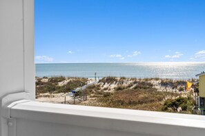A beach view with a sandy path, a flag, and a building in the distance. - 5228 E Beach Dr, perfect for 18 guests with a pool and dog-friendly amenities. (Oak Island)