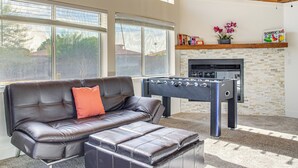 Ein modernes Wohnzimmer mit Ledersofa, Hocker und einem Kicker. Ein Kamin und ein Regal mit Büchern und Blumen sind ebenfalls vorhanden.