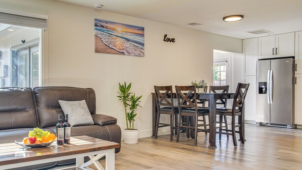 Ein modernes Wohnzimmer mit Ledersofa, Esstisch mit Stühlen, Couchtisch mit Obst und Getränken, einem Blumentopf und einer Küche mit Kühlschrank.