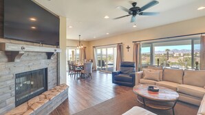 A spacious living room with a fireplace, a large TV, a ceiling fan, and a view of the mountains through the windows. - Enjoy Million-dollar Views! Lake Havasu City Home (Lake Havasu City)