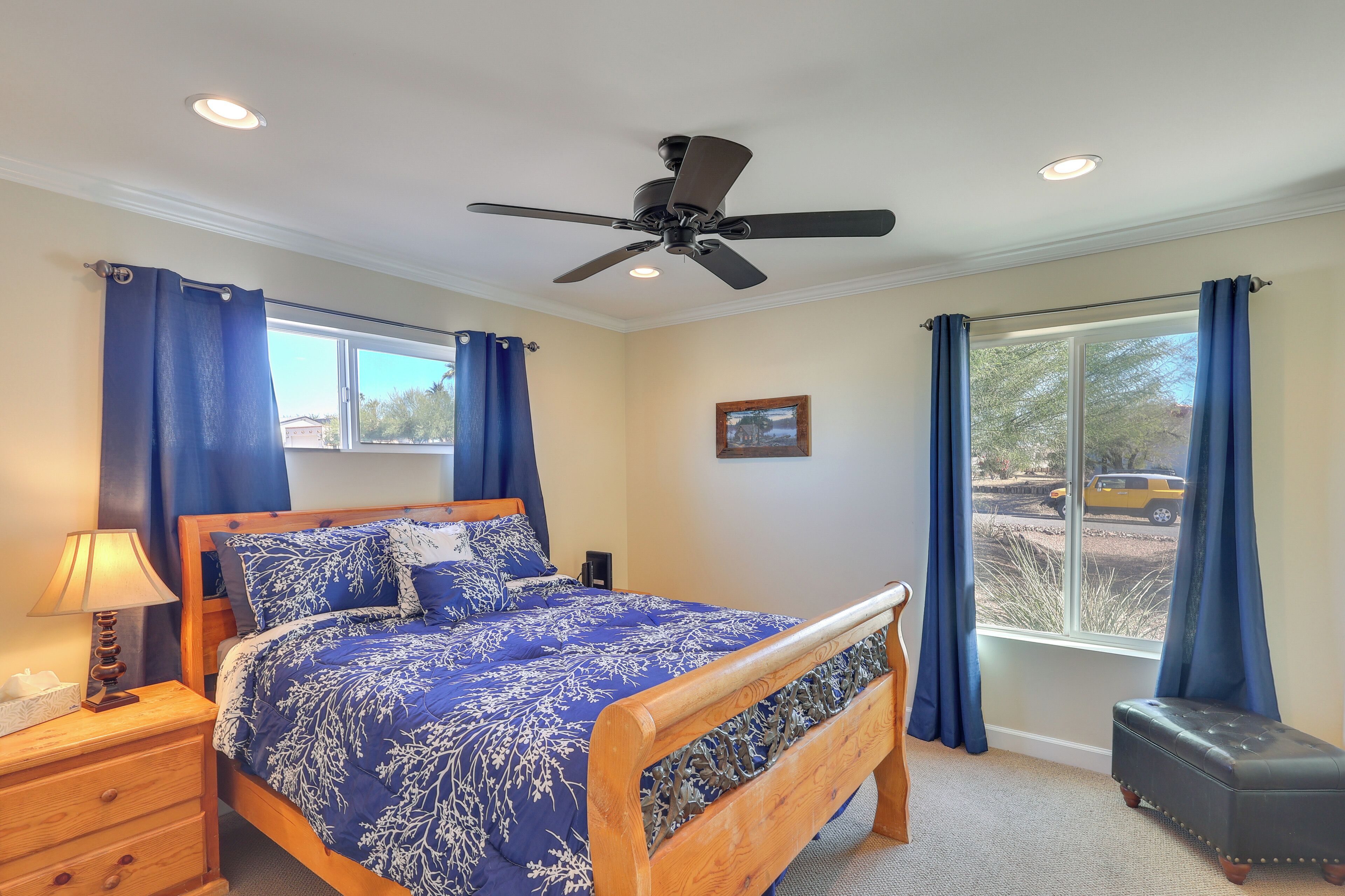 Un dormitorio con cama, mesita de noche, lámpara, ventilador de techo, ventana con cortinas y vistas al exterior.