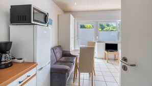 Une cuisine compacte équipée d’un four à micro-ondes, d’un réfrigérateur et d’une machine à café. On y trouve un canapé gris et une petite table à manger avec des chaises. Un téléviseur est posé sur un meuble en bois à l’arrière-plan.