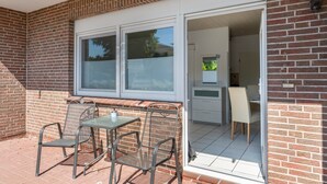 Une terrasse avec une table et des chaises, une porte-fenêtre coulissante donnant sur un espace intérieur comprenant une cuisine et un coin repas.