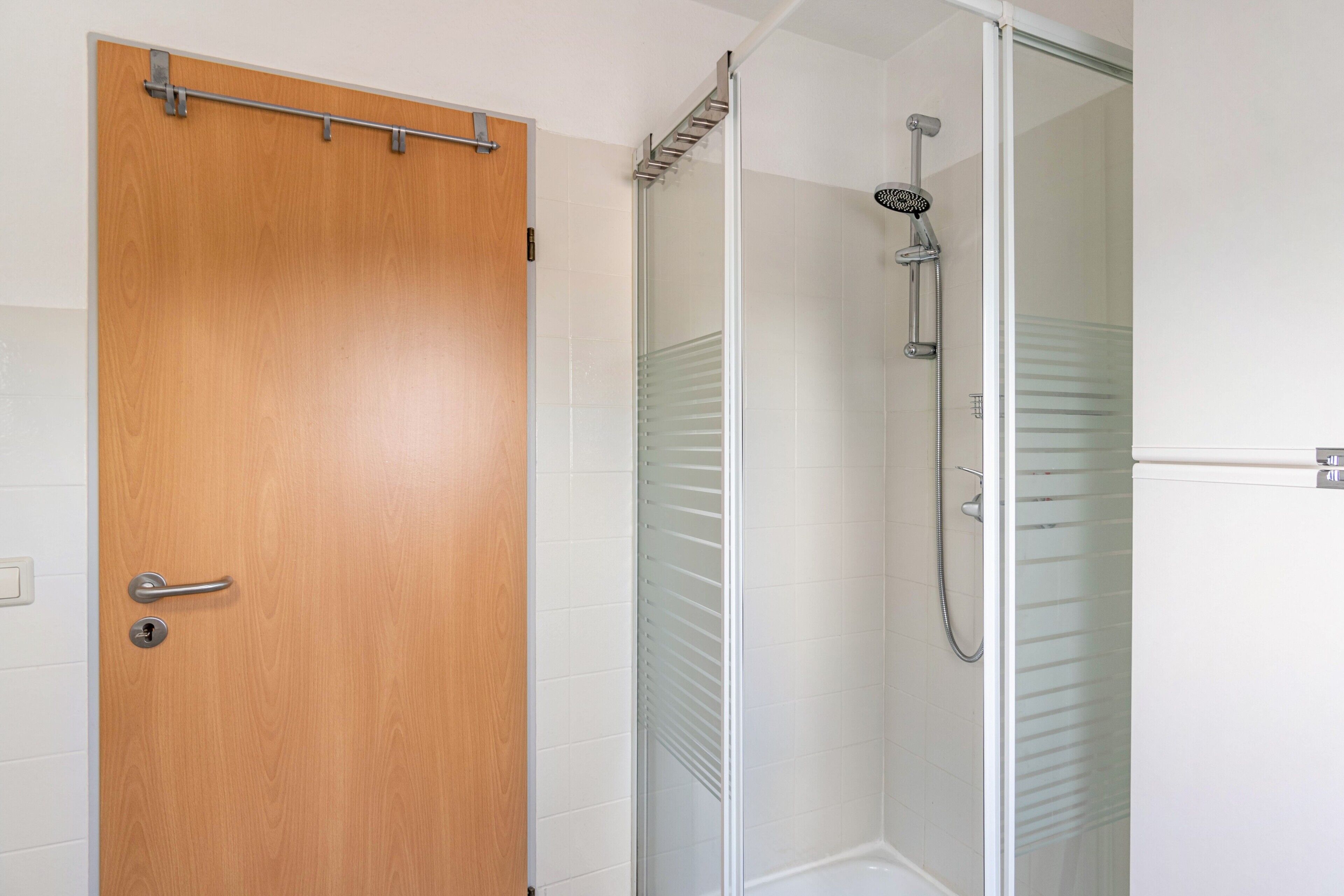 A bathroom with a wooden door, a glass shower enclosure, and white tiled walls.