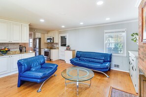 Een ruime woonkamer met een keuken, twee blauwe leren banken, een glazen salontafel en een groot raam dat voor natuurlijk licht zorgt.