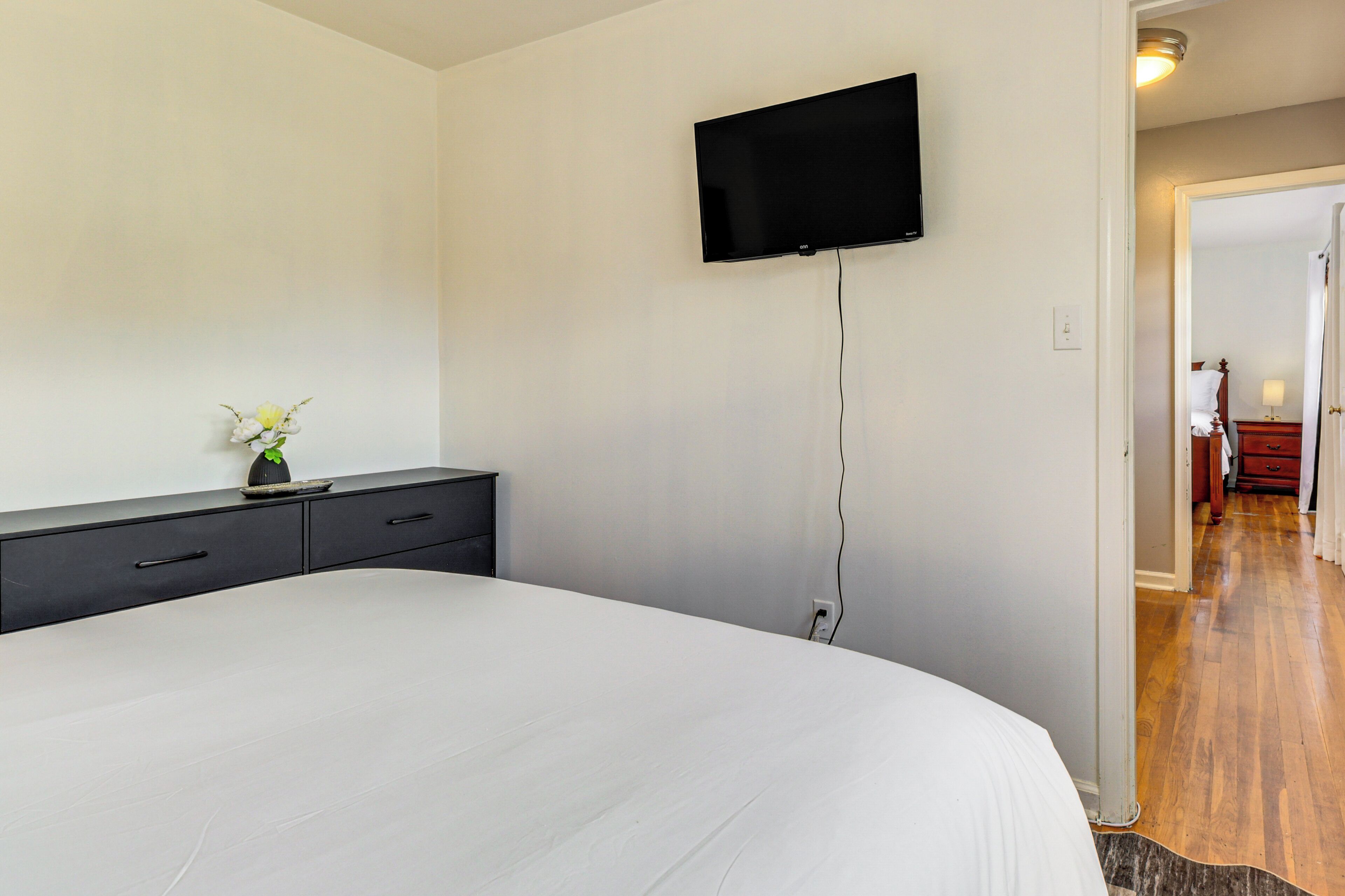 A bedroom with a large bed, a flat-screen TV mounted on the wall, a small bedside table with a vase of flowers, and a wooden floor.