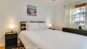 Una cama bien tendida con sábanas blancas, dos mesitas de noche con lámparas, un cuadro en la pared y una ventana con cortinas.