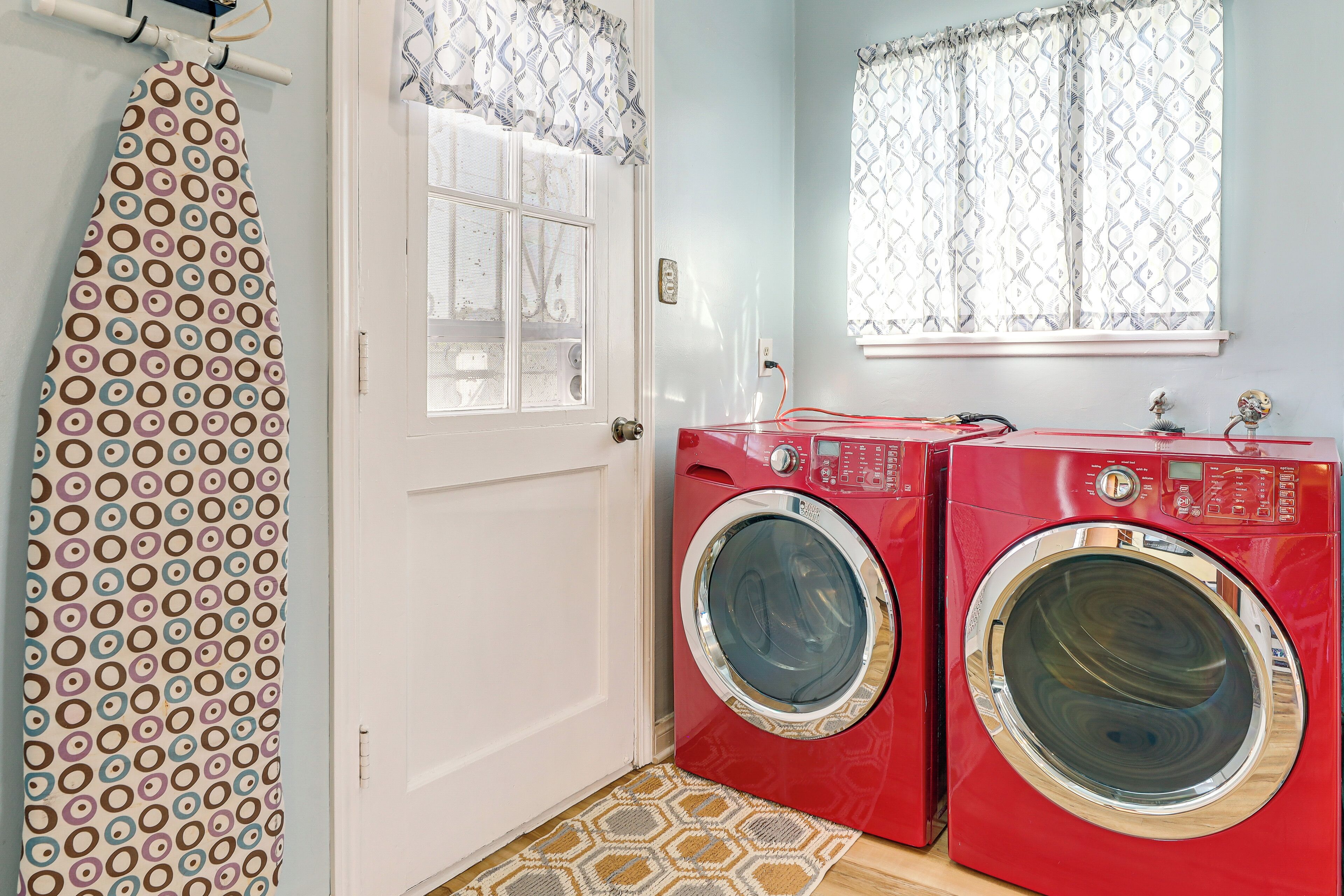 A laundry room with two red washing machines, a patterned shower curtain, and a window with patterned curtains.