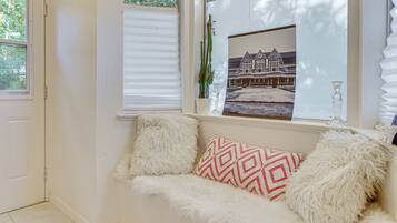 A cozy room with a white sofa, fluffy pillows, a framed picture, and a window with blinds.
